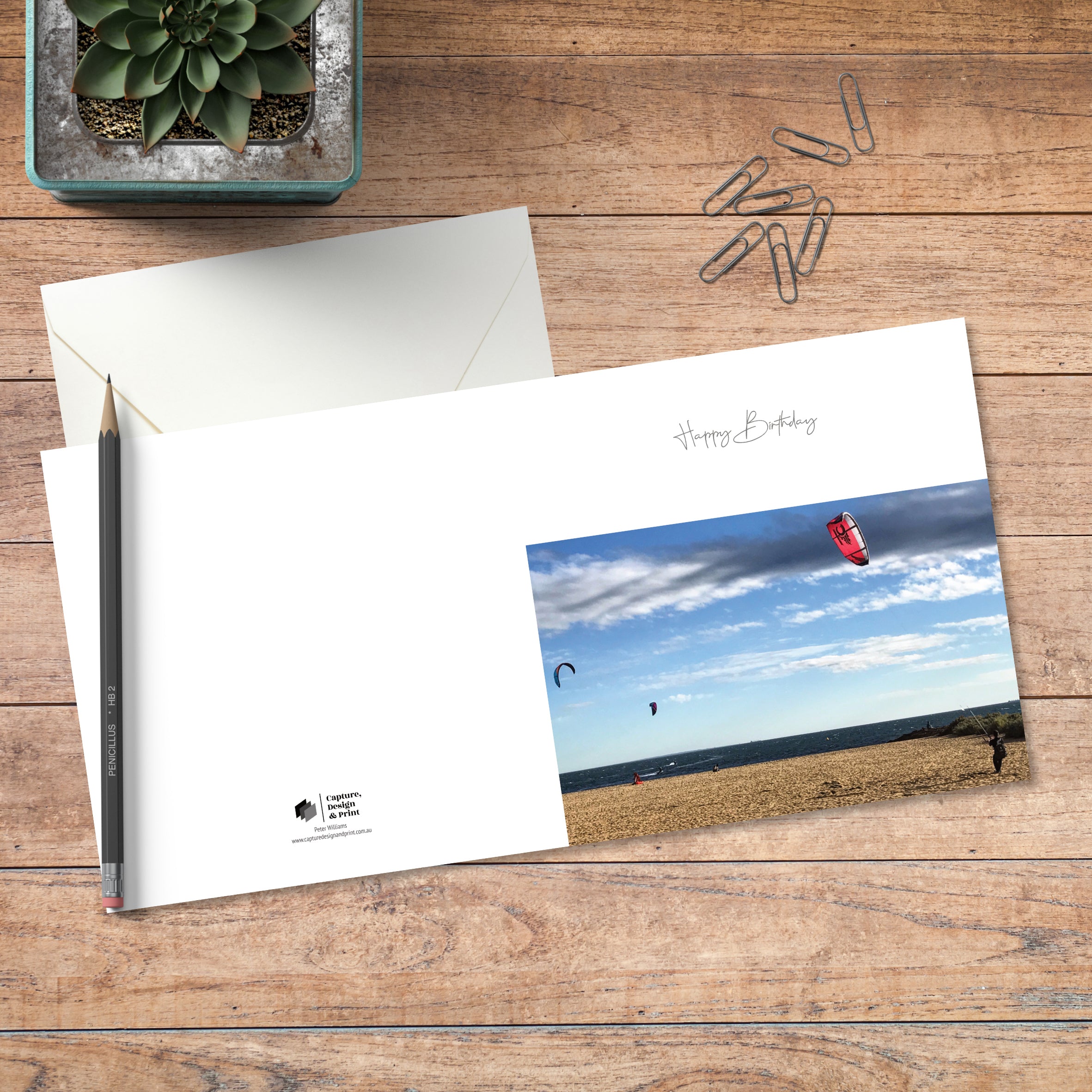 A small square birthday card in colour with full spread displayed on timber, featuring kite surfers at the beach on a winter’s day