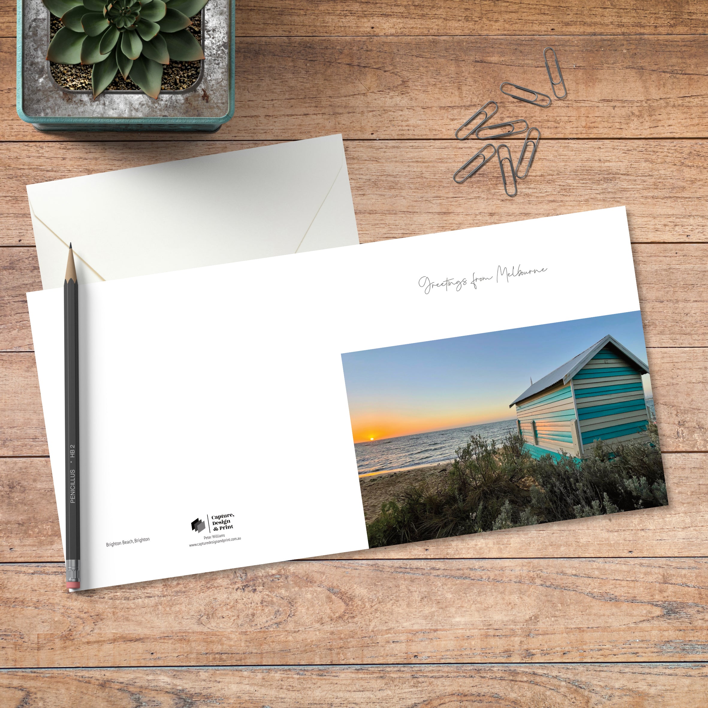 A small square greeting card in colour with full spread displayed on timber, featuring a Brighton bathing box at sunset