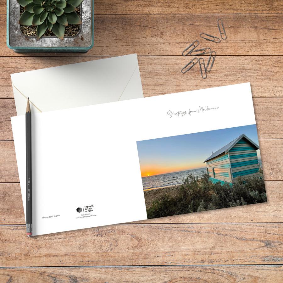 A small square greeting card in colour with full spread displayed on timber, featuring a Brighton bathing box at sunset