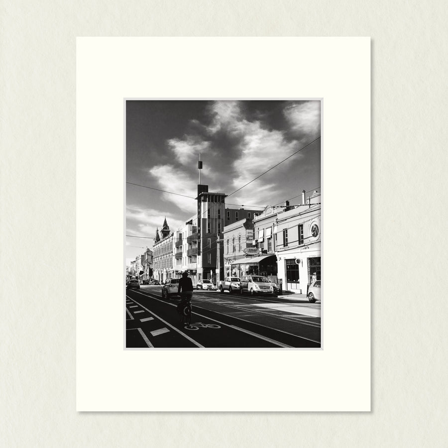 Ready to Frame Print: Fitzroy - a frame mount containing a B&W print of an urban street, classic buildings, cyclist and dramatic sky