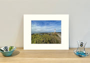 Frame mount containing a colour print of of a lighthouse with grassland and a stormy sky sitting upright on sideboard