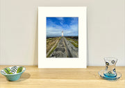 Frame mount containing a colour print of of a lighthouse at the end of a bluestone path sitting upright on sideboard