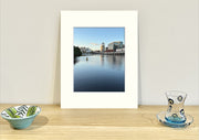 Frame mount containing a colour print of a beautiful river with reflections of old buildings in the distance sitting upright on sideboard