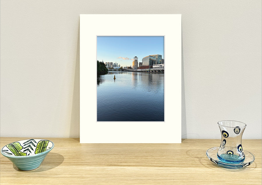 Frame mount containing a colour print of a beautiful river with reflections of old buildings in the distance sitting upright on sideboard