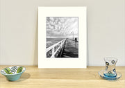 Frame mount containing a B&W print of dramatic storm clouds and fisherman sitting upright on sideboard with objets d’art to the sides