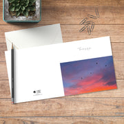 A small square greeting card in colour with full spread displayed on timber, featuring birds silhouetted against a sunset sky