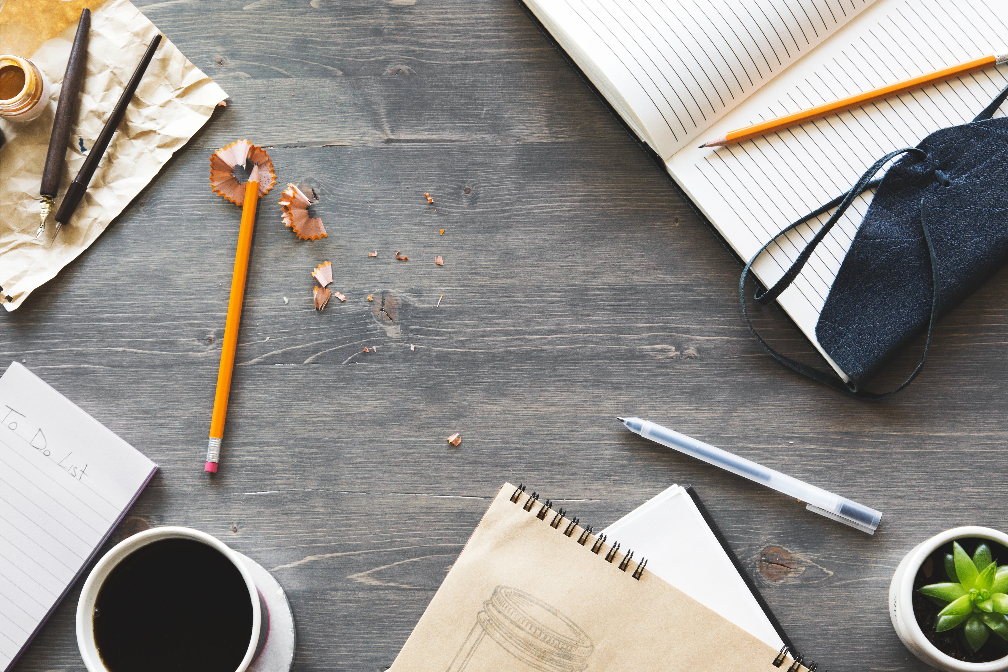 A flatlay of notebook, notepad, pencil with pencil shavings,, pen, cup of coffee and tiny pot plant